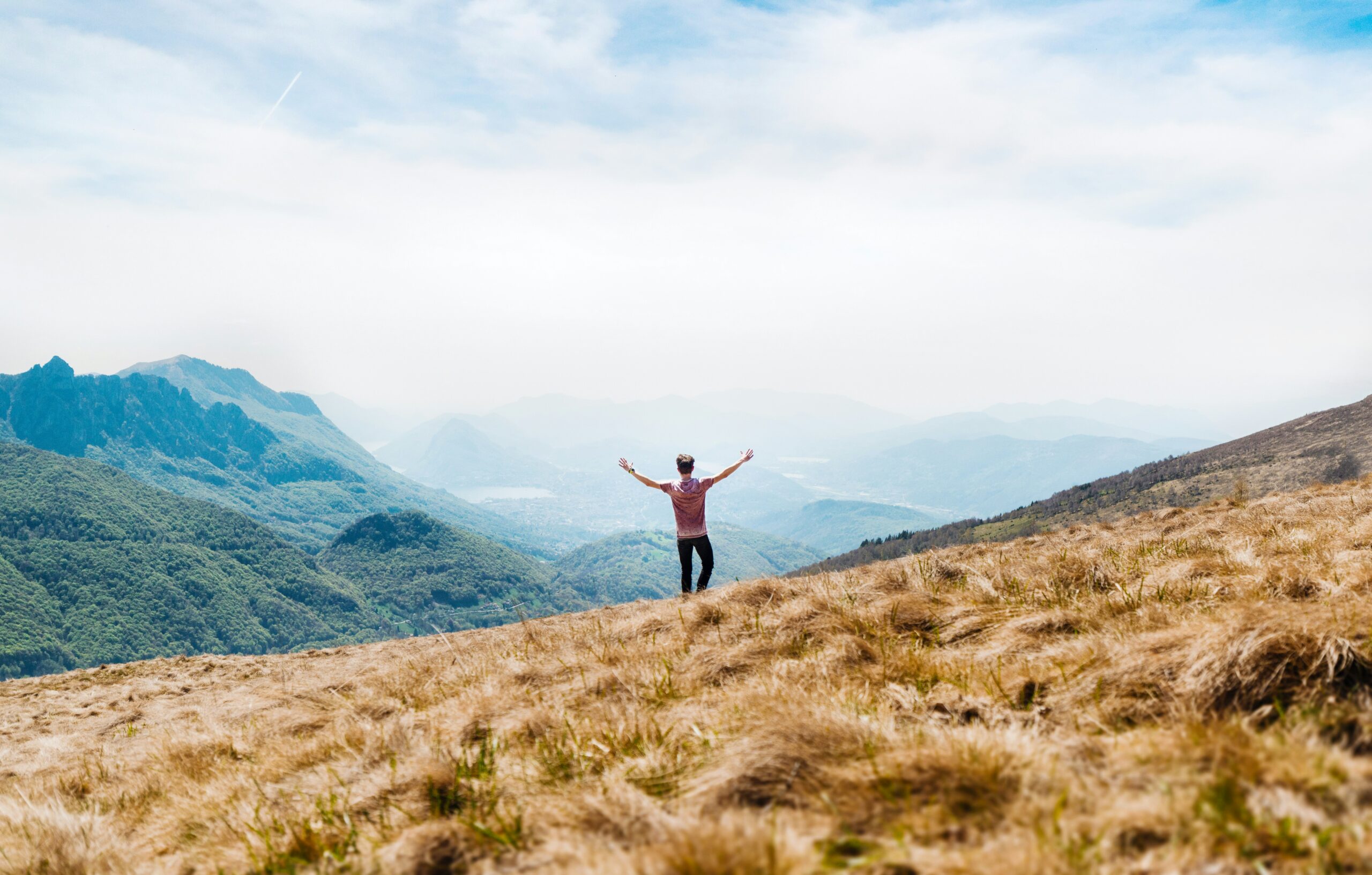 Man standing on a grassy mountain ridge with arms outstretched, overlooking vast layers of misty green hills and valleys under a blue sky.